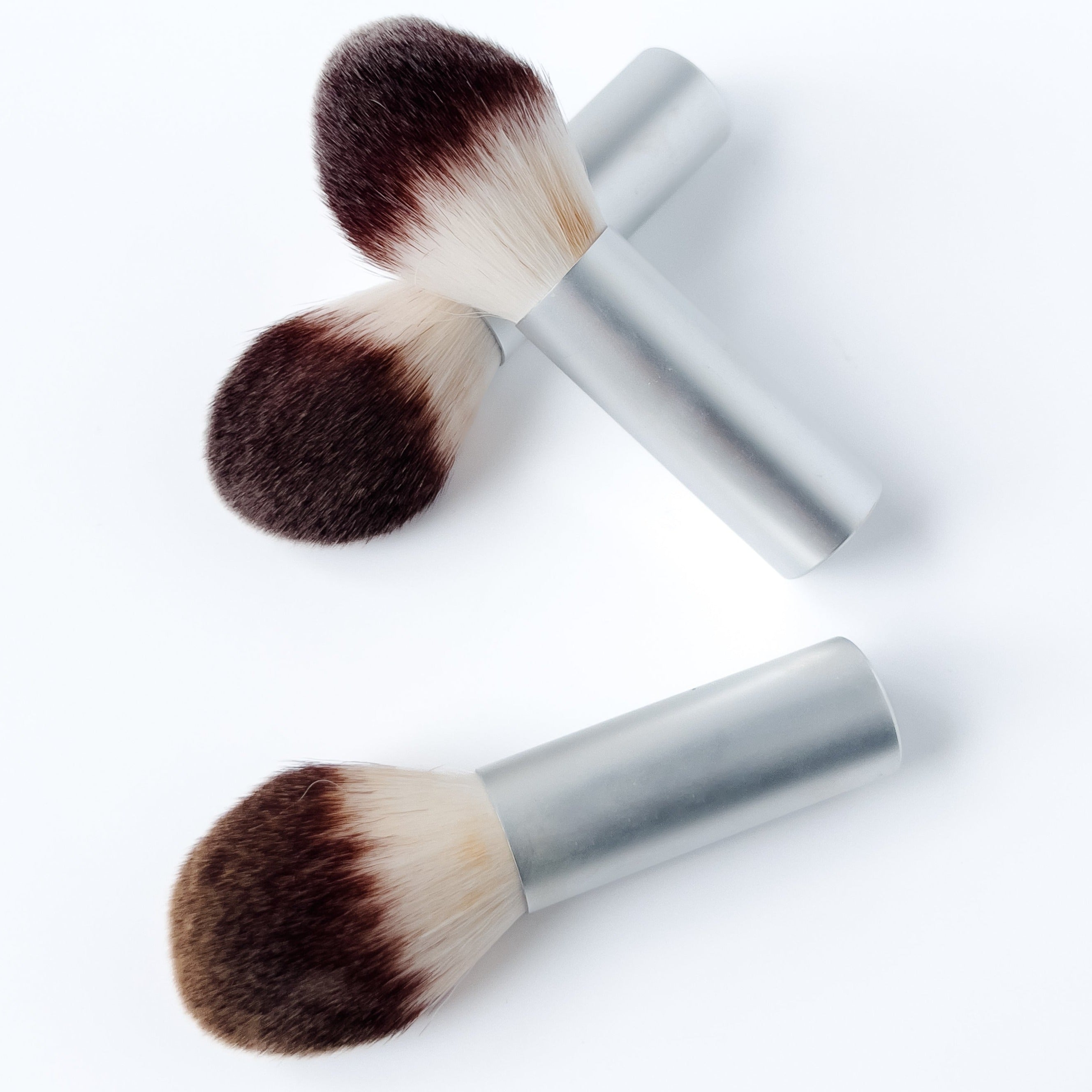 Three large foundation brushes are shown scattered on a white background. The brush has a silver body with "La Bella Donna" in white. The bristles start white and become a deep brown towards the tips.